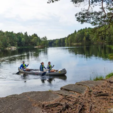 Tre personer paddlar kanot på en stilla sjö omgivet av grönskande skog. Populär utflykt i Göteborgs natur.